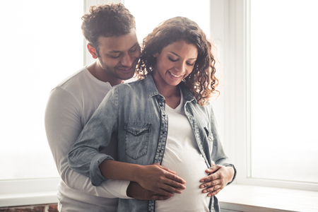 Handsome Afro American man and his beautiful pregnant wife are hugging and smiling while standing near the window at homeの写真素材