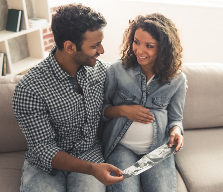 Handsome Afro American man and his beautiful pregnant wife are watching sonogram and smiling while spending time together at homeの写真素材
