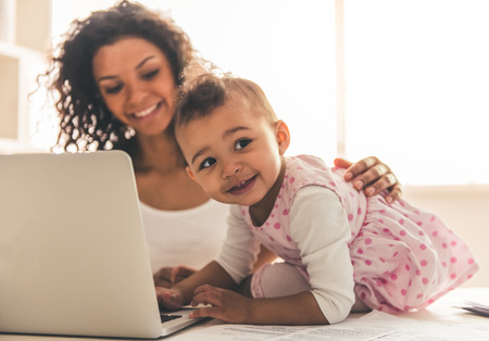 Beautiful young Afro American woman and her cute baby are using a laptop and smiling while spending time together at homeの写真素材