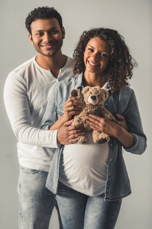 Handsome Afro American man and his beautiful pregnant wife are hugging and smiling, on gray backgroundの写真素材