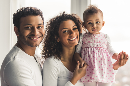 Beautiful young Afro American parents and their cute baby are looking at camera and smiling while standing near the window at homeの写真素材