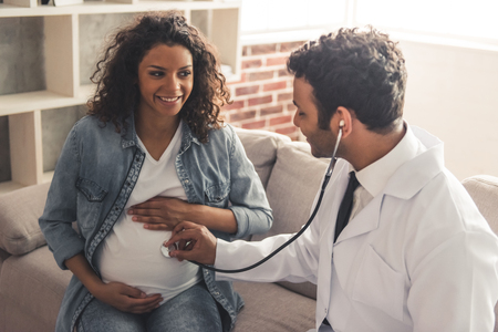 Handsome Afro American doctor in white coat is consulting beautiful pregnant woman in his officeの写真素材