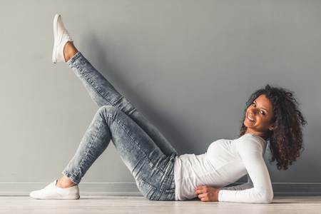 Beautiful young Afro American woman is looking at camera and smiling while sitting on the floorの写真素材