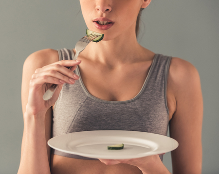 Eating disorder. Cropped image of girl holding a plate with a slice of cucumber on it, isolated on whiteの写真素材