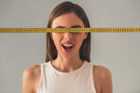 Suffering from anorexia. Portrait of girl with a tape measure in front of her eyes, on gray...