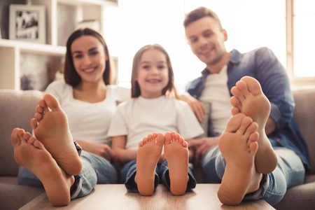 Charming little girl and her beautiful young parents are looking at camera and smiling while sitting on sofa at home. Bare feet in focusの写真素材