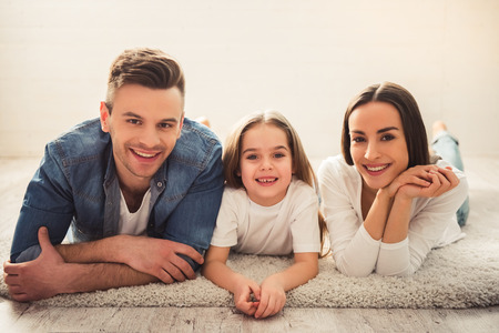 Charming little girl and her beautiful young parents are looking at camera and smiling while lying on the floor at homeの写真素材