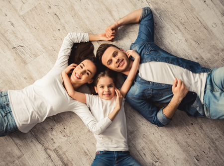 Top view of cute little girl and her beautiful young parents looking at camera and smiling while lying on the floor at homeの写真素材