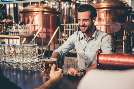 Handsome young waiter is giving beer to clients and smiling while working in pubの写真素材