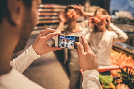 Handsome dad is taking photo of his beautiful cheerful wife and daughter while they are doing shopping in the supermarketの写真素材