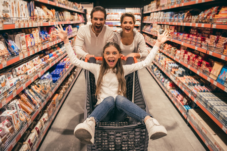 Beautiful couple and their daughter are having fun while doing shopping in supermarket. Girl is sitting in trolleyの写真素材