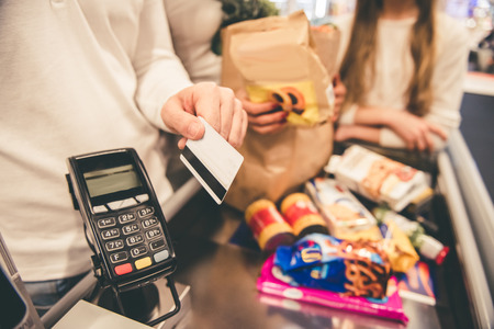 Cropped image of beautiful parents and their daughter standing at the cash desk in supermarket. Dad is using a credit cardの写真素材