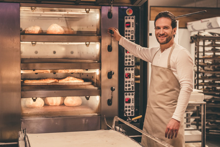 Handsome supermarket worker is looking at camera and smiling while working with oven at the bakeryの写真素材