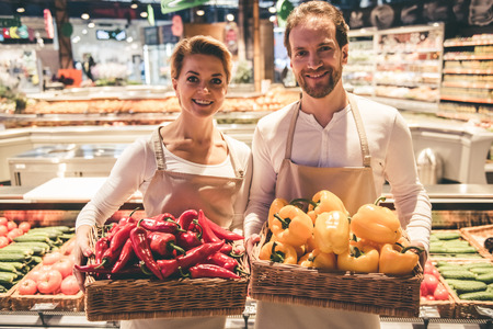 Beautiful couple of supermarket workers are holding vegetables, looking at camera and smilingの写真素材