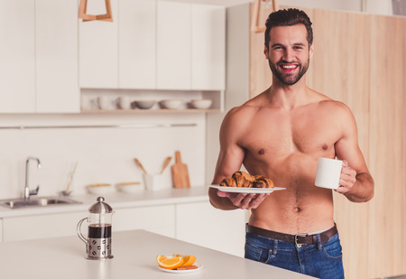 Handsome shirtless young man is holding croissants and coffee, looking at camera and smiling while having breakfast in kitchenの写真素材