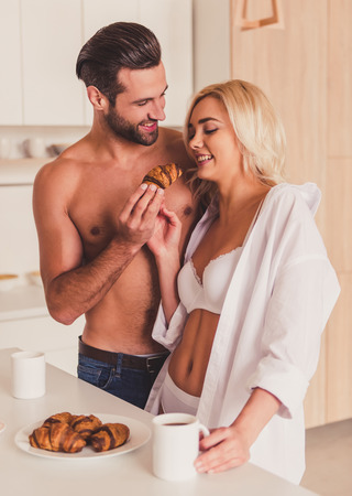 Beautiful young half-naked couple is eating croissants, drinking coffee and smiling while having a breakfast in kitchen in the morningの写真素材