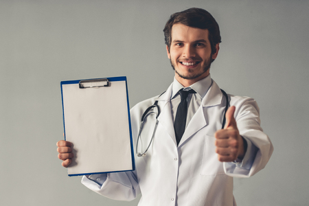 Handsome young doctor in white coat is holding a folder, showing Ok sign, looking at camera and smiling, on gray backgroundの写真素材