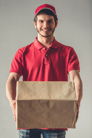 Handsome young delivery worker in red uniform is holding a cardboard box, looking at camera and smiling, on gray backgroundの写真素材