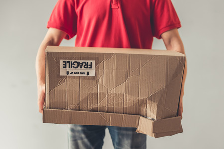 Cropped image of handsome young delivery worker in red uniform holding a cardboard box, on gray backgroundの写真素材