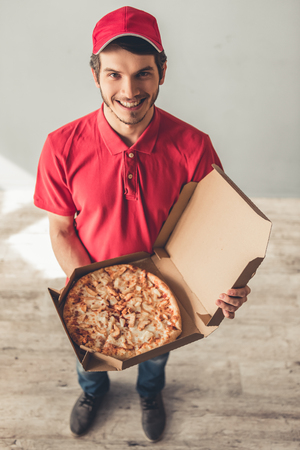Handsome young delivery worker in red uniform is holding a pizza, looking at camera and smiling, on gray backgroundの写真素材