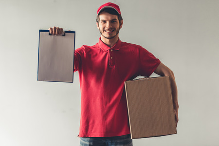 Handsome young delivery worker in red uniform is holding a cardboard box and document, looking at...