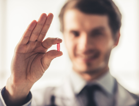 Handsome young doctor in white coat is holding a pill, hand with medicine in focusの写真素材