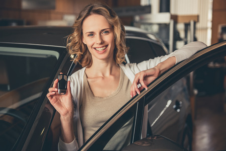Beautiful young woman is holding a key, looking at camera and smiling while standing near the new car in car dealershipの写真素材