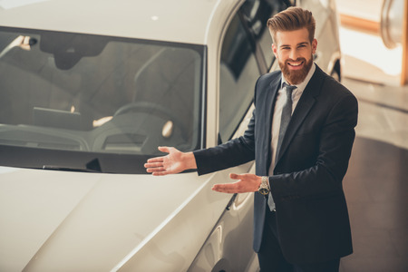 Handsome bearded businessman in suit is pointing on the car, looking at camera and smiling while standing in car dealershipの写真素材