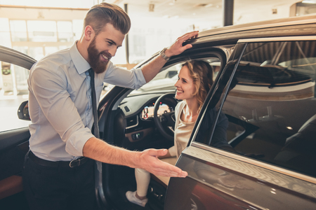 Beautiful young woman is talking to handsome bearded sales manager while choosing a car in dealershipの写真素材
