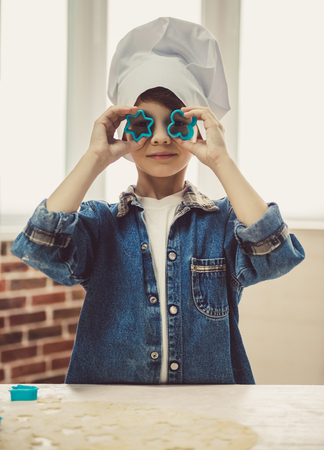 Cute little boy in chef hat is playing with cookie cutters while making cookies in kitchenの写真素材