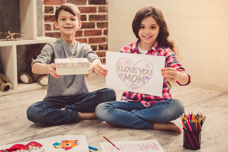 Cute children are showing their handmade presents for mom and smiling while sitting on the floor at homeの写真素材