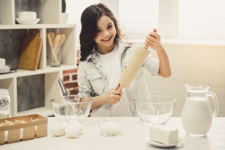 Cute little girl is holding a rolling pin, looking at camera and smiling while baking in the kitchenの写真素材