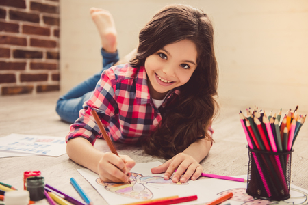 Cute little girl is drawing, looking at camera and smiling while lying on the floor at homeの写真素材