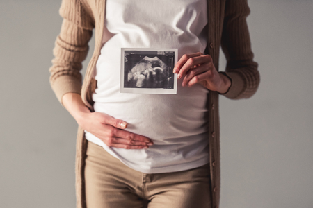 Cropped image of beautiful pregnant woman stroking her tummy and holding a sonogram, on gray backgroundの写真素材