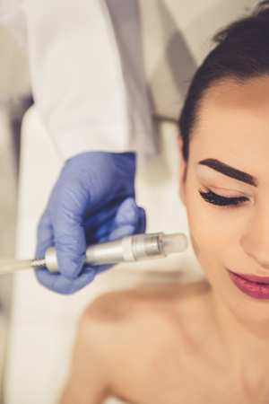 Beautiful young woman is getting face skin treatment. Doctor in medical gloves is undertaking the procedure using modern equipmentの写真素材