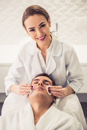 Handsome man is getting face skin treatment. Beautiful cosmetician is smiling while clearing patient's faceの写真素材