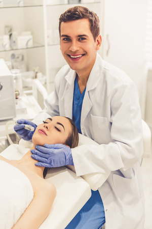 Beautiful young woman is getting face skin treatment. Handsome doctor in medical gloves is undertaking the procedure using modern equipmentの写真素材