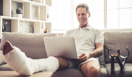 Handsome mature man with broken leg in gypsum is using a laptop and smiling while sitting on sofa at homeの写真素材