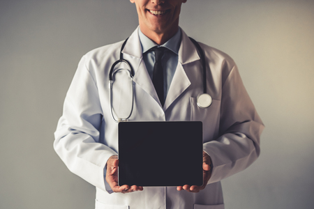 Cropped image of handsome mature doctor in white coat holding a digital tablet, on gray backgroundの写真素材
