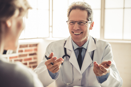 Handsome mature doctor in white coat is talking to the patient and smiling during the consultation in his officeの写真素材