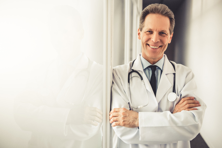 Handsome mature doctor in white coat is looking at camera and smiling while standing with crossed arms in hospital corridorの写真素材