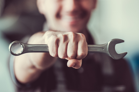 Handsome mechanic in uniform is holding a spanner and smiling while standing in auto serviceの写真素材