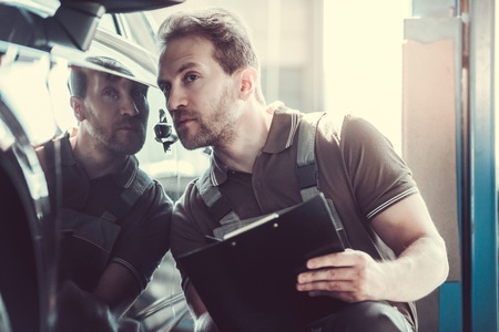 Handsome mechanic in uniform is making notes examining car in auto serviceの写真素材