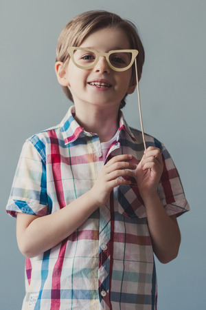 Funny little boy is holding paper glasses on wooden stick, looking at camera and smiling, on gray backgroundの写真素材