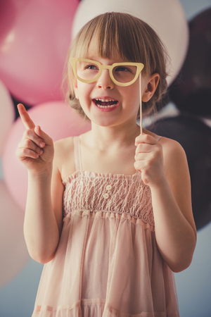 Cute little princess in dress is posing with paper eyeglasses and smiling, on colorful balloons backgroundの写真素材