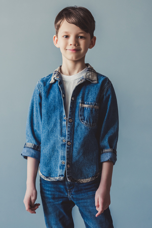 Handsome little boy in jean clothes is looking at camera and smiling, on gray backgroundの写真素材