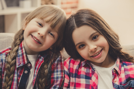 Charming little girls are looking at camera and smiling while sitting on couch at homeの写真素材