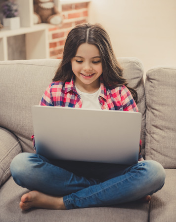 Cute little girl in casual clothes is using a laptop and smiling while sitting on sofa at homeの写真素材