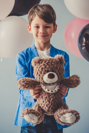 Handsome little boy in stylish clothes is holding a teddy bear, looking at camera and smiling, on gray background with colorful balloonsの写真素材