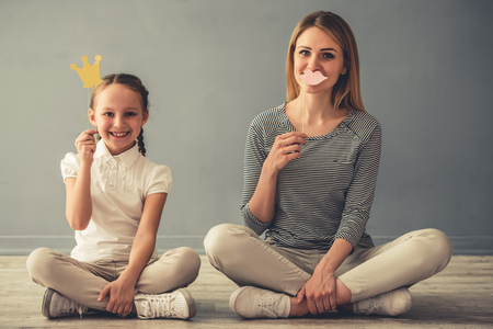 Beautiful woman and her cute little daughter are holding paper crown and lips, looking at camera and smiling while sitting on the floorの写真素材